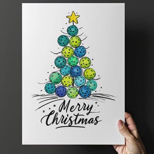 May include: A white print featuring a Christmas tree made of colourful pickleballs, topped with a yellow star. The words "Merry Christmas" are written in elegant script below the tree. The print is held by a hand.