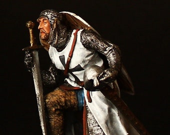 Puede incluir: Una figurita miniatura pintada a mano de un caballero arrodillado con armadura blanca y una cruz roja en el pecho. Sostiene una espada y un casco. La figurita está colocada sobre una base marrón.