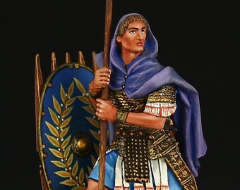 May include: A painted miniature figurine of a Roman soldier wearing a purple cloak and holding a spear with a golden eagle on top. The soldier is standing on a green base with a brown rock and a golden helmet.