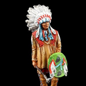 Puede incluir: Una figurita pintada a mano de un guerrero nativo americano con un tocado de plumas, una camisa con cuentas y sosteniendo un escudo y un hacha. La figurita está de pie sobre una base marrón.