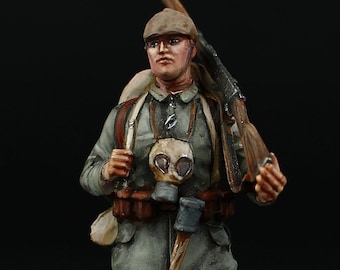May include: A painted miniature figurine of a soldier wearing a gas mask and carrying a rifle. The soldier is wearing a brown hat, a grey uniform, and brown boots. The figurine is standing on a green base.