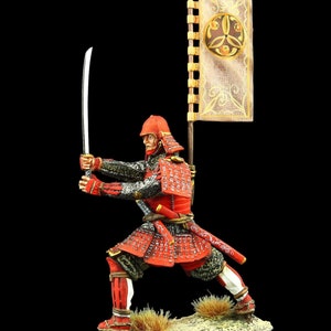 Puede incluir: Una figurita pintada de rojo y dorado de un guerrero samurái en pose de lucha, sosteniendo una espada y una bandera con un diseño dorado. La figurita está de pie sobre una base marrón con hierba verde.