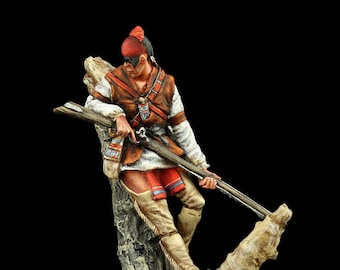 May include: A painted miniature figure of a Native American warrior in a red and white feathered headdress, holding a rifle and perched on a tree branch. The figure is wearing a brown leather vest, a red and blue sash, and white leggings. The tree branch is painted in shades of brown and green, and the base is a square piece of brown material with green and brown foliage.