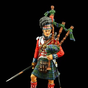 May include: A painted figurine of a Scottish piper wearing a kilt, a red jacket, and a white shirt. He is holding a bagpipe and a sword. The figurine is standing on a small base.