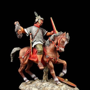 Könnte beinhalten: Eine bemalte Figur eines römischen Soldaten zu Pferd, der in seiner linken Hand einen abgetrennten Kopf hält. Der Soldat trägt eine Kettenhemdtunika, einen roten Umhang und einen Helm mit einer Feder. Das Pferd ist braun mit weißen Abzeichen und bäumt sich auf seinen Hinterbeinen auf. Die Figur steht auf einem felsigen Sockel.