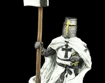 May include: A miniature figurine of a knight in white armor holding a spear with a white and black flag. The knight is wearing a white robe with a black cross on the chest. The figurine is standing on a brown base.