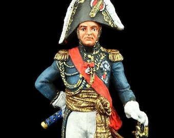 May include: A painted figurine of a military officer wearing a blue uniform with gold trim, a white sash, and a black bicorne hat with white feathers. The officer is holding a sword and standing on a round base.