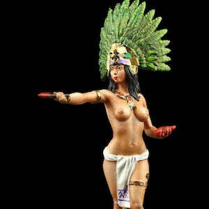May include: A painted figurine of a woman wearing a green feathered headdress and a white skirt. She is holding a red knife in her right hand and a red object in her left hand. She is standing on a brown base with a red and orange design.