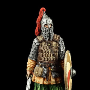 May include: A hand-painted metal figurine of a warrior wearing chainmail armour, a helmet with a red plume, and holding a sword and shield. The figurine is standing on a brown base.