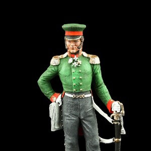 May include: A painted figurine of a military officer wearing a green uniform with gold trim, a red sash, and a black hat with a red band. The officer is holding a sword and standing on a brown base.