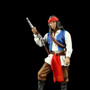 Puede incluir: Figura de un guerrero nativo americano, de pie sobre una roca. Viste una cinta roja en la cabeza, camisa blanca, chaleco azul, pantalones marrones y botas negras. Sostiene un rifle. Una faja roja cuelga de su cinturón.