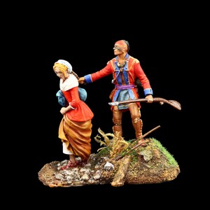 May include: A painted figurine set depicting a Native American man in a red and blue coat holding a rifle and leading a woman in a red and brown dress.