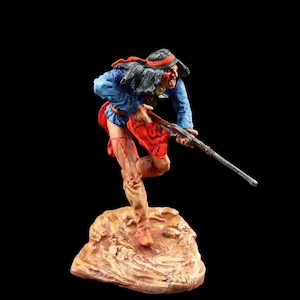 Może przedstawiać: Ręcznie malowana figurka indiańskiego wojownika w czerwono-niebieskim stroju, trzymającego karabin i stojącego na brązowej podstawie.