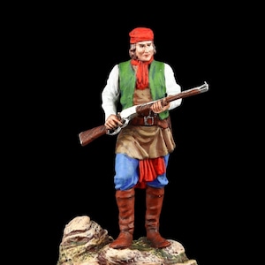 Op de afbeelding: Een beeldje van een grensman met een geweer. De figuur draagt een rode hoed en sjaal, een groen vest en een bruin schort over een blauwe broek. Bruine laarzen maken de outfit compleet. De figuur staat op een rotsachtige basis tegen een zwarte achtergrond.