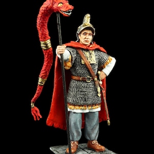 May include: A painted figurine of a Roman soldier wearing a red cloak and holding a spear with a red dragon head. The soldier is wearing a chainmail tunic and a helmet with a gold crest.