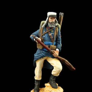 May include: A painted figurine of a soldier wearing a blue uniform, white hat, and carrying a rifle. The soldier is standing on a rocky base.