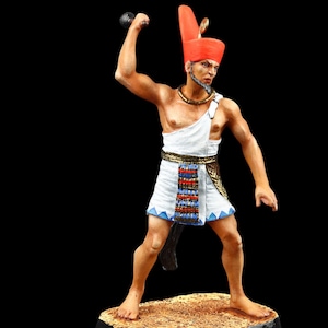 Könnte beinhalten: Eine bemalte Figur eines alten ägyptischen Kriegers, der einen roten und goldenen Kopfschmuck, einen weißen Kilt mit blauem und goldenem Besatz und einen goldenen Gürtel trägt. Der Krieger hält einen schwarzen Knüppel in seiner rechten Hand und steht auf einem braunen Sockel.