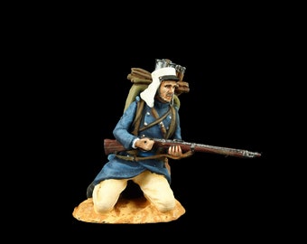 May include: A painted figurine of a soldier kneeling and holding a rifle. He is wearing a blue uniform, a white head scarf, and a brown backpack. The figurine is set on a brown base.