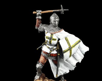 Puede incluir: Figura detallada de un caballero medieval con armadura completa, sosteniendo un arma en alto. El caballero viste una túnica y capa blancas con un emblema de cruz negra y amarilla. La figura se alza sobre una pequeña base texturizada sobre fondo negro.