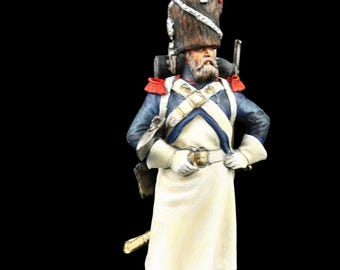 May include: A miniature figurine of a soldier in a blue coat, white apron, and tall hat with a red plume. The figure has a white beard and is standing on a small, rectangular base. The background is black.