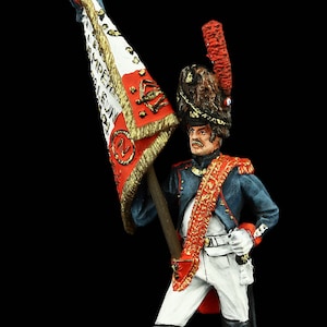 Op de afbeelding: Een geschilderd miniatuurbeeldje van een Franse soldaat die een vlag vasthoudt met de inscriptie "LIBERTE EGALITE FRATERNITE". De soldaat draagt een blauw uniform met rode en gouden accenten, een witte hoed met een zwarte bontkraag en zwarte laarzen. De vlag is rood, wit en blauw met gouden versieringen.