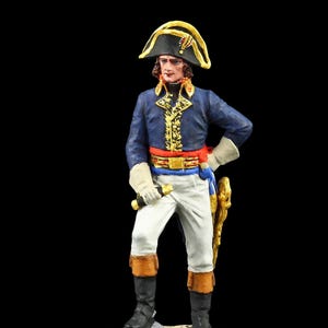 Może przedstawiać: Miniaturowa figurka postaci historycznej w niebieskiej kurtce ze złotymi wykończeniami, białych spodniach i czarnym kapeluszu. Figurka stoi na małej, teksturowanej podstawie, trzymając miecz. Figurka jest przedmiotem kolekcjonerskim.