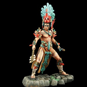 Puede incluir: Una figurita pintada a mano de un guerrero Maya con un tocado de plumas, una armadura intrincada y un taparrabos. El guerrero sostiene un arma en una mano y un escudo en la otra. La figurita está de pie sobre una base de piedra.