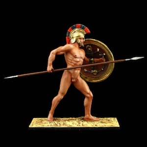 Può includere: Una statuetta dipinta di un guerriero greco con un elmo rosso e nero e che tiene una lancia e uno scudo. Il guerriero è nudo e ha uno scudo dorato con un disegno in bianco e nero.