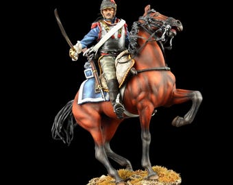 May include: A detailed figurine of a cavalry soldier in a blue uniform with a silver breastplate and helmet, holding a curved sword while mounted on a rearing brown horse. The horse is positioned on a textured, rocky base with patches of dry grass.