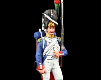 May include: A miniature figurine of a soldier in a blue coat, white pants, and a tall black hat. The soldier is holding a rifle and standing on a small, rectangular base. The figure has a red and green plume on the hat and a red sash.