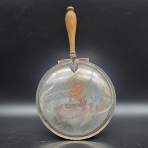 May include: A silver-toned, round crumb catcher with a wooden handle. The crumb catcher features a copper-colored lion emblem and a decorative edge. The handle is light brown and attached with a silver-toned hinge.