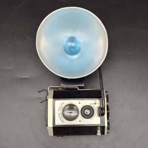 May include: Vintage Kodak Brownie Reflex camera with a large, light blue flash reflector. The camera is black and silver with a round lens and the text "Brownie Reflex" and "Eastman Kodak Co." printed on the front.