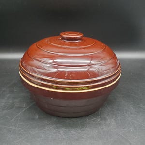 Puede incluir: Una fuente de cerámica marrón con tapa. La fuente tiene forma redonda con una tapa que tiene un pequeño asa. La fuente tiene un acabado brillante y una banda de color claro en el medio. La fuente es adecuada para cocinar y servir.