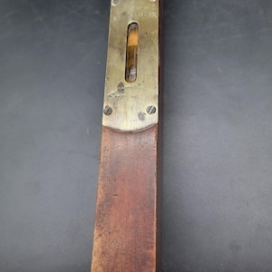 Op de afbeelding: Een vintage houten waterpas met een messing bovenkant. De messing bovenkant heeft een waterpas en is vastgezet met twee schroeven. Het hout is donkerbruin van kleur en de waterpas lijkt goed gebruikt.