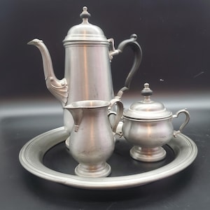 Op de afbeelding: Een vintage tinnen theeservies, bestaande uit een hoge koffiepot, een melkkan, een suikerpot en een serveerblad. De koffiepot heeft een zwart handvat en een decoratieve tuit. De set wordt gepresenteerd op een zwart oppervlak.