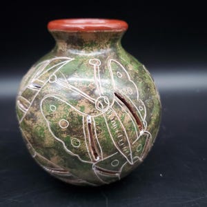 May include: A small, round ceramic vase with a green and brown mottled finish. The vase features white etched butterfly and leaf designs. The rim is a reddish-brown color. The vase is a decorative item.