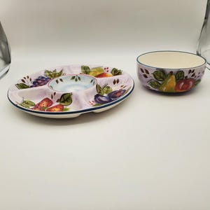 May include: A ceramic serving set featuring a divided oval platter and a matching bowl. The set is painted with colorful fruit illustrations, including grapes, strawberries, and plums, on a light purple background. The rims are trimmed with a dark blue border.