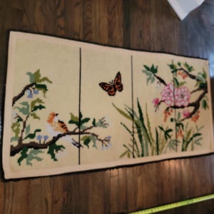 May include: A rectangular, cream-colored needlepoint rug with a nature scene. The rug features a bird perched on a branch, a butterfly, and pink flowers. The border is black.