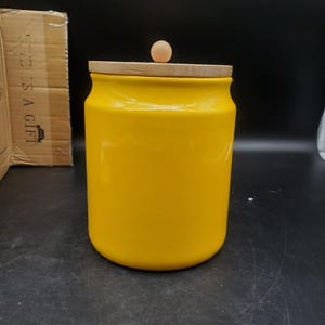 May include: A bright yellow cylindrical ceramic storage canister with a smooth, glossy finish. The container features a flat, light-colored wooden lid topped with a small, round, light-colored wooden knob handle centered on the top surface.