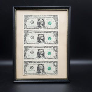 May include: A framed display featuring four one-dollar bills. Each bill displays George Washington's portrait and the text "The United States of America." The bills are green and black, set within a cream-coloured mount, in a black frame.