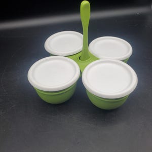 Puede incluir: Un servidor de condimentos verde lima con cuatro cuencos redondos y tapas blancas. Los cuencos están conectados por una base central verde lima que incluye un asa vertical alta para transportar. Cada tapa blanca tiene una textura circular.