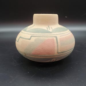 Puede incluir: Un pequeño jarrón redondo de cerámica beige con cuello corto. El jarrón está decorado con patrones geométricos en tonos rosa, verde y gris. El diseño incluye triángulos y formas rectangulares.