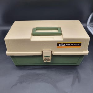 Può includere: Una scatola da pesca Plano bicolore. La parte superiore è beige e la parte inferiore verde. Ha una maniglia verde e un fermaglio in metallo. Il logo Plano è sul davanti in nero, arancione e bianco.