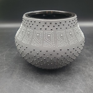 May include: A matte charcoal gray ceramic bowl with a wide, rounded body and a black interior. The exterior surface is decorated with a repeating pattern of raised black and white dots arranged in vertical rectangular columns and rows.