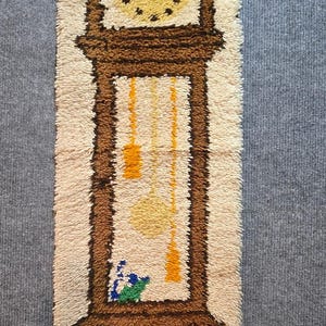May include: A rectangular latch hook wall hanging featuring a tall brown grandfather clock design on a cream background. The clock face is yellow with black markings, and the interior shows yellow pendulums, blue flowers, and an orange cat.