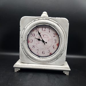 May include: White, square-shaped table clock with a distressed finish. The clock face features Roman numerals and the words "Victoria Station London 1865". The clock sits on a matching rectangular base and has a small decorative top.