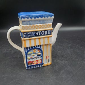 Puede incluir: Una tetera de cerámica con forma de edificio, techo azul y toldo de rayas amarillas. El frente presenta un escaparate con queso y botellas de vino, con el texto WINE & CHEESE STORE impreso en azul sobre un fondo de color blanco.