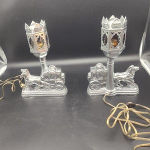 May include: A pair of silver-toned metal lamps featuring a horse-drawn carriage base and a lantern-style top with cut-out heart details. Each lamp includes an electrical cord with a switch, designed to hold a single light bulb inside the lantern.