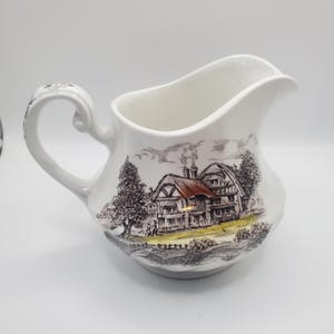 Puede incluir: Jarra de cerámica blanca con asa y boquilla curvadas. La jarra presenta una detallada ilustración en negro y marrón de una escena rural con una casa, árboles y figuras. El asa tiene un diseño decorativo.