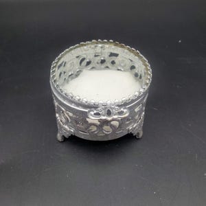 May include: A small, round, silver-colored jewelry box with ornate filigree detailing. The box has a clear, glass-like top and a soft, white interior. The box is sitting on a black surface.
